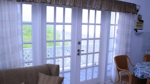 Ein Zimmer mit einer großen Schiebeglasdoor zum Balkon, ein Rattansofa, ein Esstisch mit Stühlen und eine kleine Wandregal mit Gegenständen.
