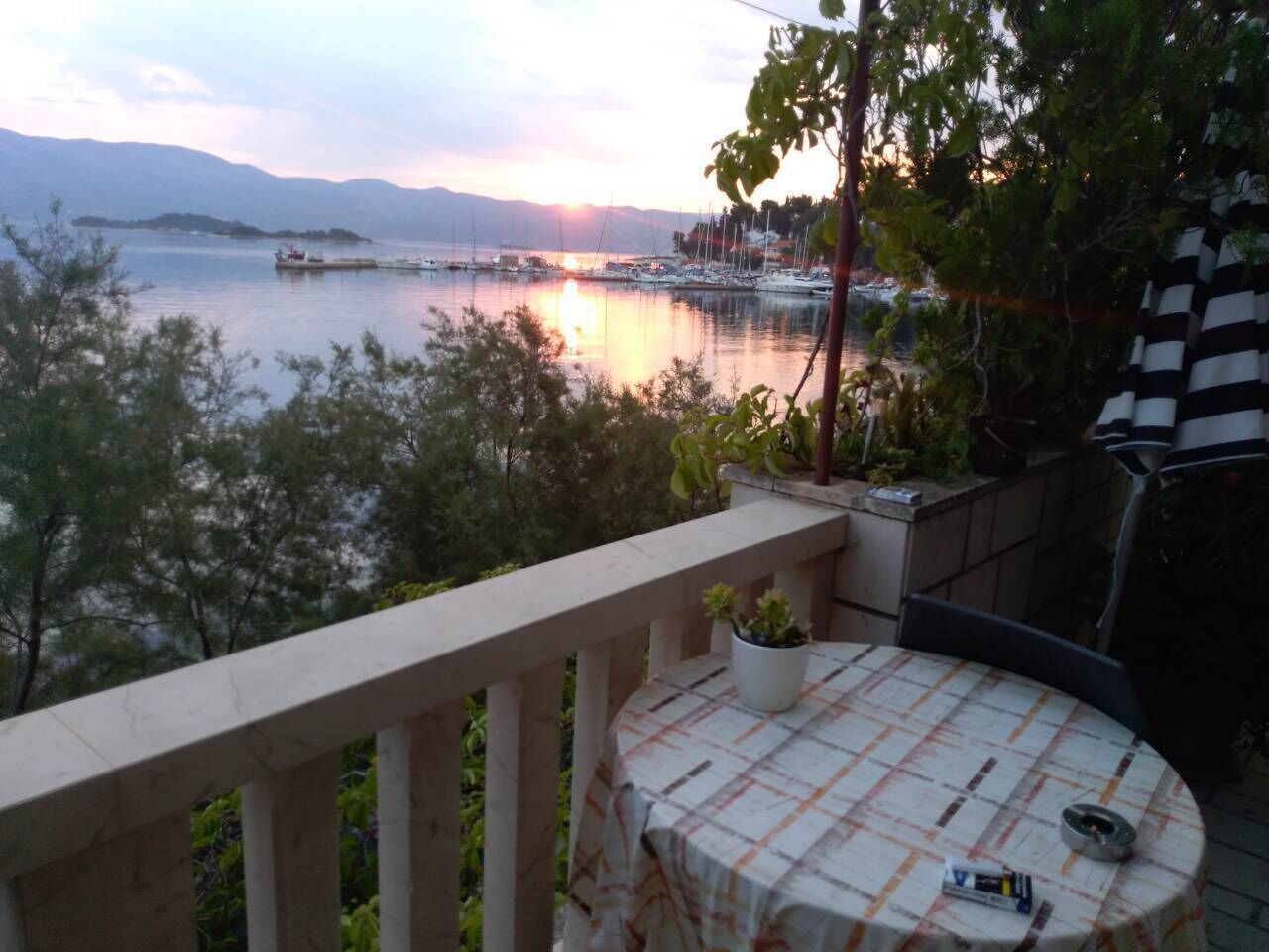 A balcony with a table and chairs, overlooking a marina with boats and a sunset.