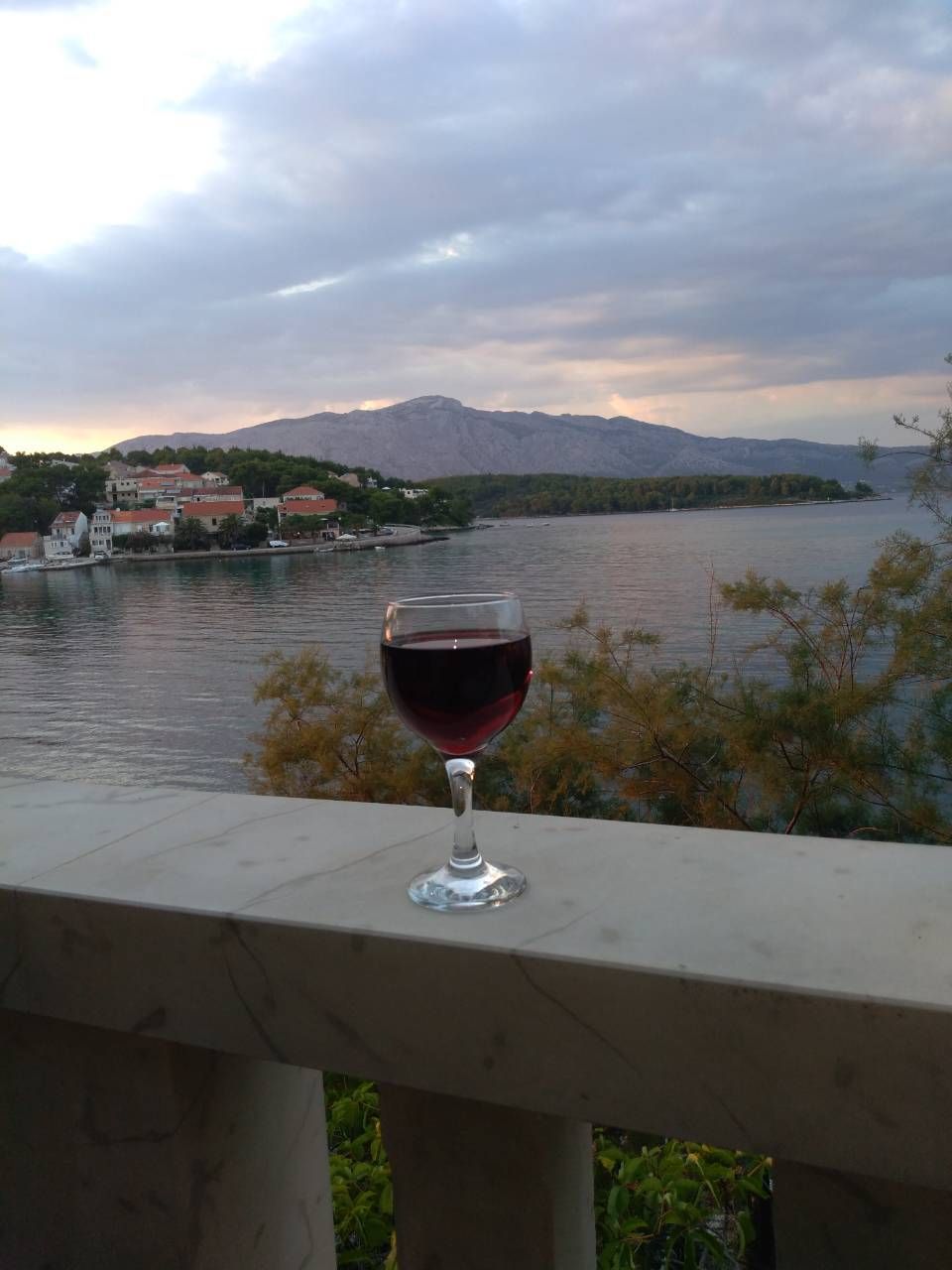 Et glas rødvin på en balkon med udsigt over en sø, med bygninger og bjerge i baggrunden.