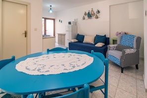 A dining area with a round blue table, chairs, and a white doily. There is a sofa with blue cushions and a patterned armchair. A refrigerator and a small table with flowers are in the background.