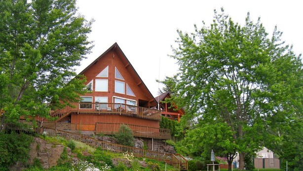 A large, multi-story wooden house with a prominent balcony on the upper floor, surrounded by lush greenery and a rocky hillside. - Premier Chalet Home on Beautiful Williams Lake - Consistently #1 in Region.  (Cheney)