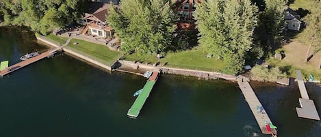 An aerial view of a lakeside property with a dock, a house with a large deck, and surrounding trees.