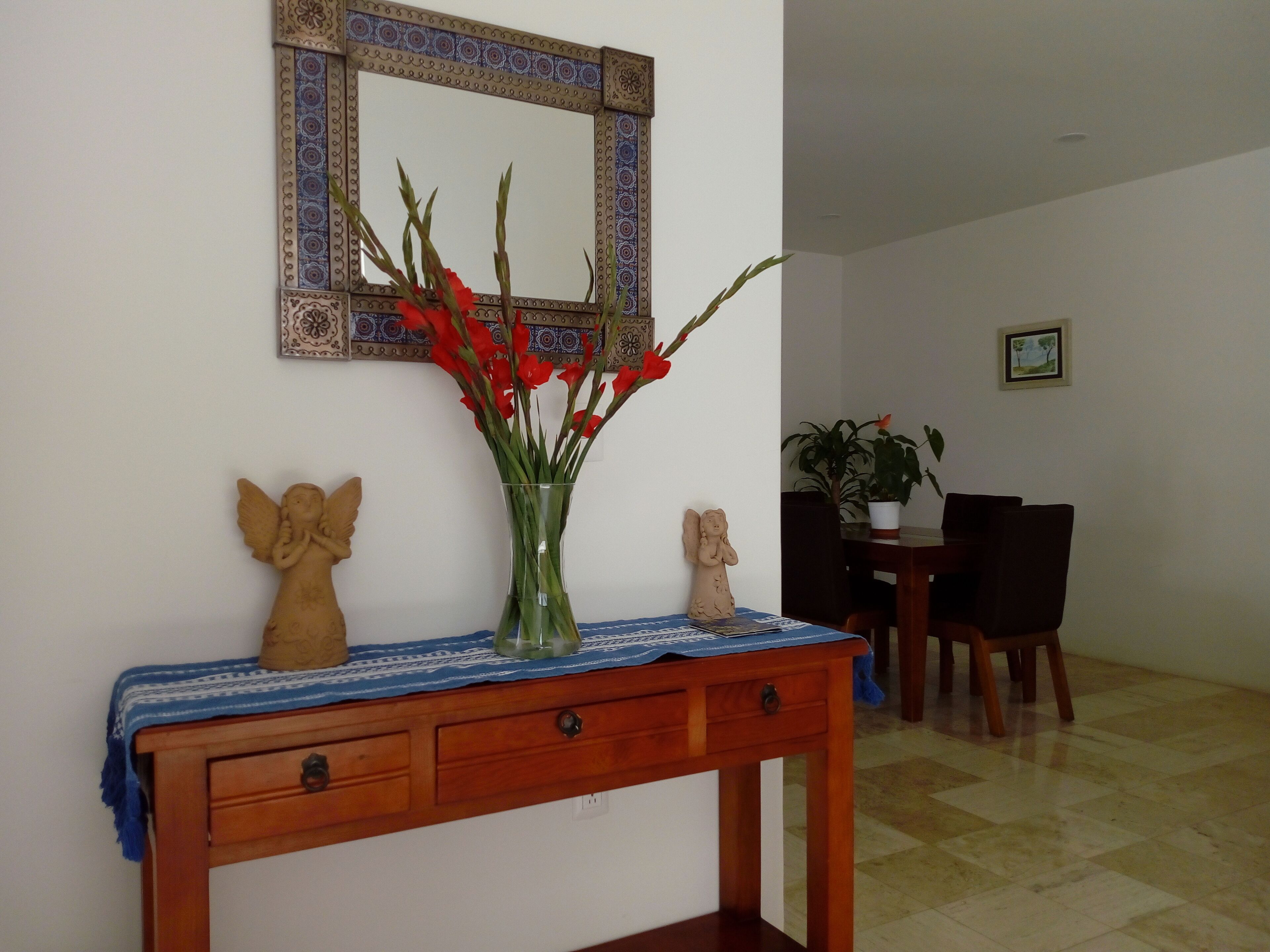 A wooden console table with a vase of red flowers, angelic sculptures, and a mirror with a decorative frame.