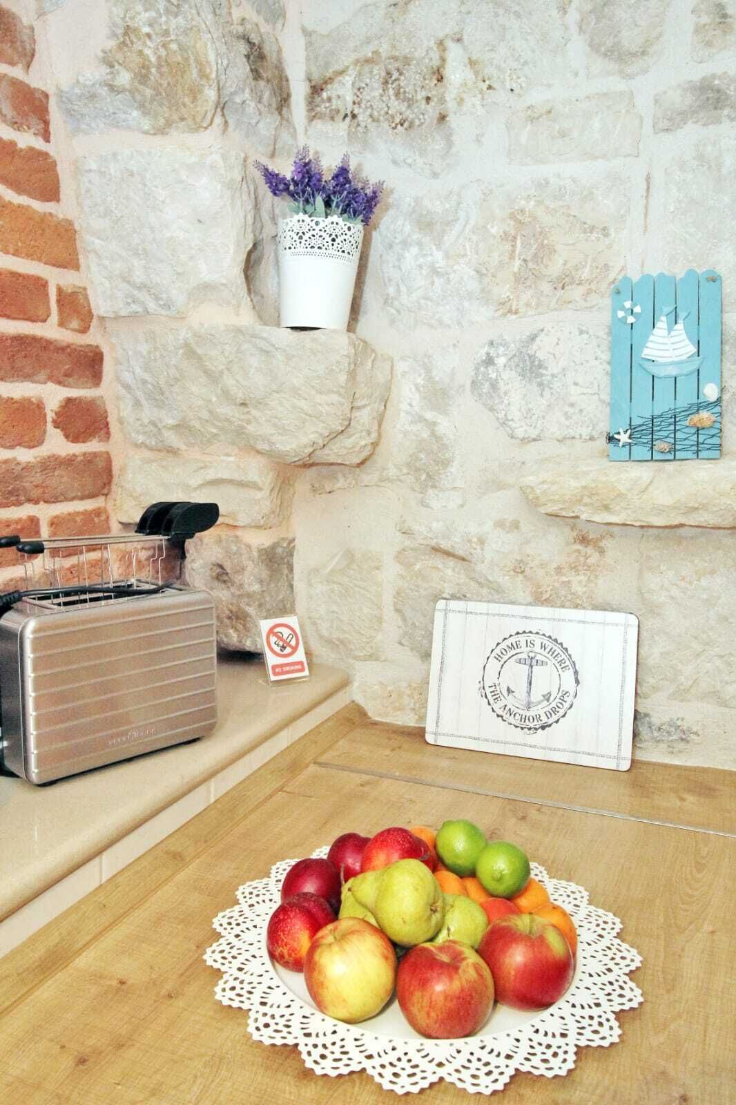 Une cuisine rustique avec un mur en pierre, une radio ancienne, un panier à fruits recouvert d’une nappe en dentelle et un élément décoratif mural.