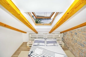 A small room with a sloped ceiling and a skylight, wooden beams, a bed with striped bedding, and a stone wall.