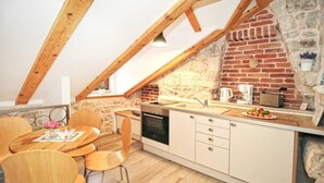 Cocina compacta con pared de piedra, vigas de madera, encimera blanca y zona de comedor con sillas de madera.