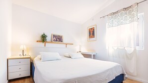 Een slaapkamer met een bed, nachtkastje, lamp en een wandplank met plant en foto.
