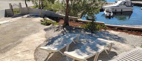 Eine Terrasse mit zwei Liegestühlen, einem kleinen Baum und einem Boot, das am Wasser liegt.