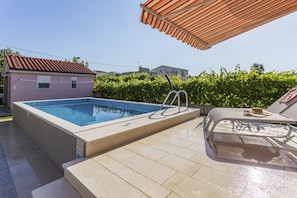 Une piscine avec une terrasse carrelée, une chaise longue et un espace abrité doté d’un auvent à rayures.
