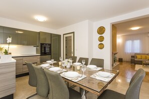 Un espace repas moderne avec une table en bois dressée pour six personnes, entourée de chaises. On aperçoit à l’arrière-plan une cuisine équipée d’un four et d’un réfrigérateur intégrés.