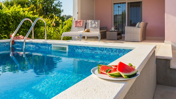 Une piscine, avec une assiette de tranches de pastèque posée sur le bord, un fauteuil de relaxation et une table sur laquelle se trouvent un chapeau et des lunettes de soleil.