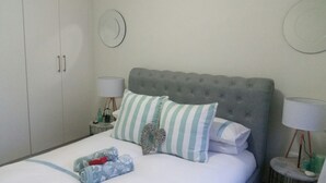 A neatly made bed with striped pillows, a grey tufted headboard, and a bedside table with a lamp and a bottle. - A Modern and Relaxed Condo in the leafy suburb of Dainfern...  (Midrand)