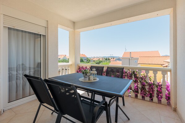 A balcony with a table and chairs, a sliding glass door, and a view of buildings and greenery.