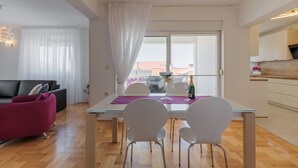 Ett modernt vardagsrum med ett matbord i glas, vita stolar, en svart soffa och en röd fåtölj. Genom fönstret syns en balkong med utsikt över byggnader.