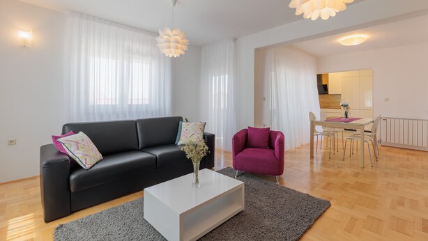 Ett modernt vardagsrum med en svart soffa, ett vitt soffbord och en lila fåtölj. Längre bak i rummet finns en matplats med ett bord och stolar.