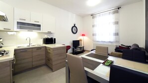 A compact kitchen with a microwave, sink, and cabinets. There is a dining table with chairs and a red lamp. A television is on a stand next to the window, which has curtains. - Cozy two bedrooms apartment in vivid neighborhood  (Zagreb)