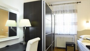 A compact hotel room with a bed, a desk with a chair, a mirror, a lamp, a radiator, and a window with sheer curtains. - Cozy two bedrooms apartment in vivid neighborhood  (Zagreb)
