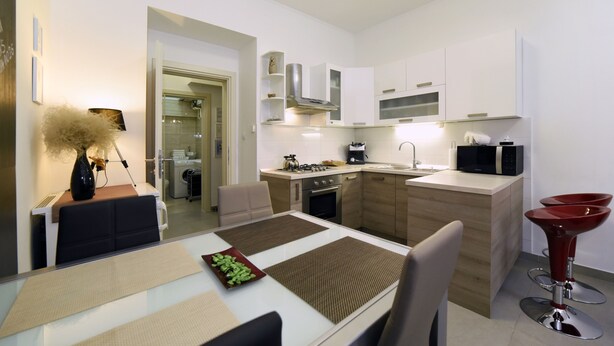 A modern kitchen with white cabinets, a gas stove, and a microwave. There is a dining area with a table and chairs, and a red bar stool. - Cozy two bedrooms apartment in vivid neighborhood  (Zagreb)