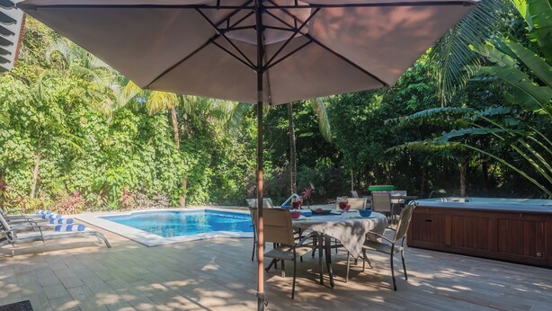 Un área junto a la piscina con mesa y sillas, una bañera de hidromasaje y un gran parasol.