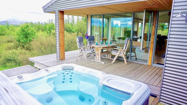 Een houten terras met een jacuzzi, buitenmeubels en een glazen eethoek met uitzicht op het omliggende landschap.