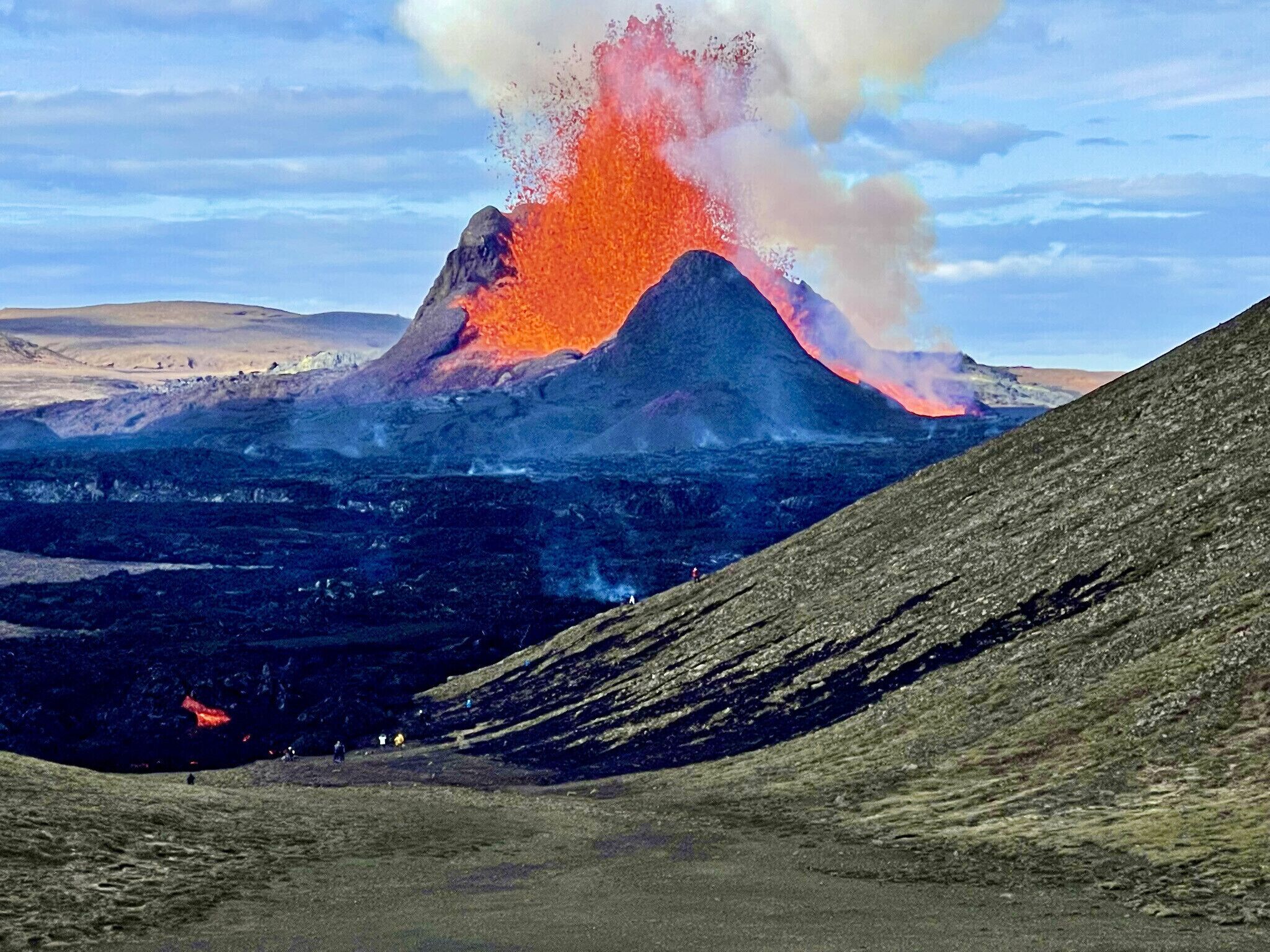Un paisaje volcánico con una erupción destacada, una zona llana con gente dispersa y una colina en primer plano.