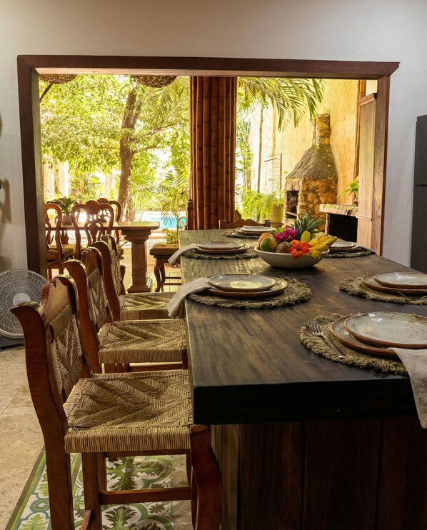 A dining area with a long wooden table set for four, surrounded by wicker chairs. A stone fireplace and a view of lush greenery are visible in the background.