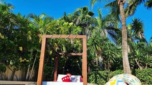 A wooden bed frame with a mattress and pillows, set against a backdrop of palm trees and greenery. - Villa Miramar Aventuras – Luxury Tropical Villa in the Mayan Riviera (Puerto Aventuras)