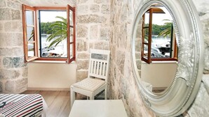 A room with a stone wall, a wooden-framed window, a white chair, a small table, and a large mirror reflecting a view of palm trees and boats.