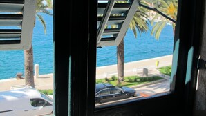 A view through a window with blinds, overlooking a coastal road with palm trees and a body of water.