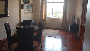 Un comedor con mesa de vidrio y sillas negras, un cuadro en la pared y un gran ventanal con cortinas.