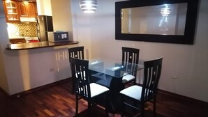 Un comedor con una mesa de vidrio y cuatro sillas, una cocina con armarios de madera y un refrigerador, y un espejo grande en la pared.