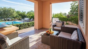 Uma área de estar coberta ao ar livre com móveis de vime, piscina e vista para o verde.
