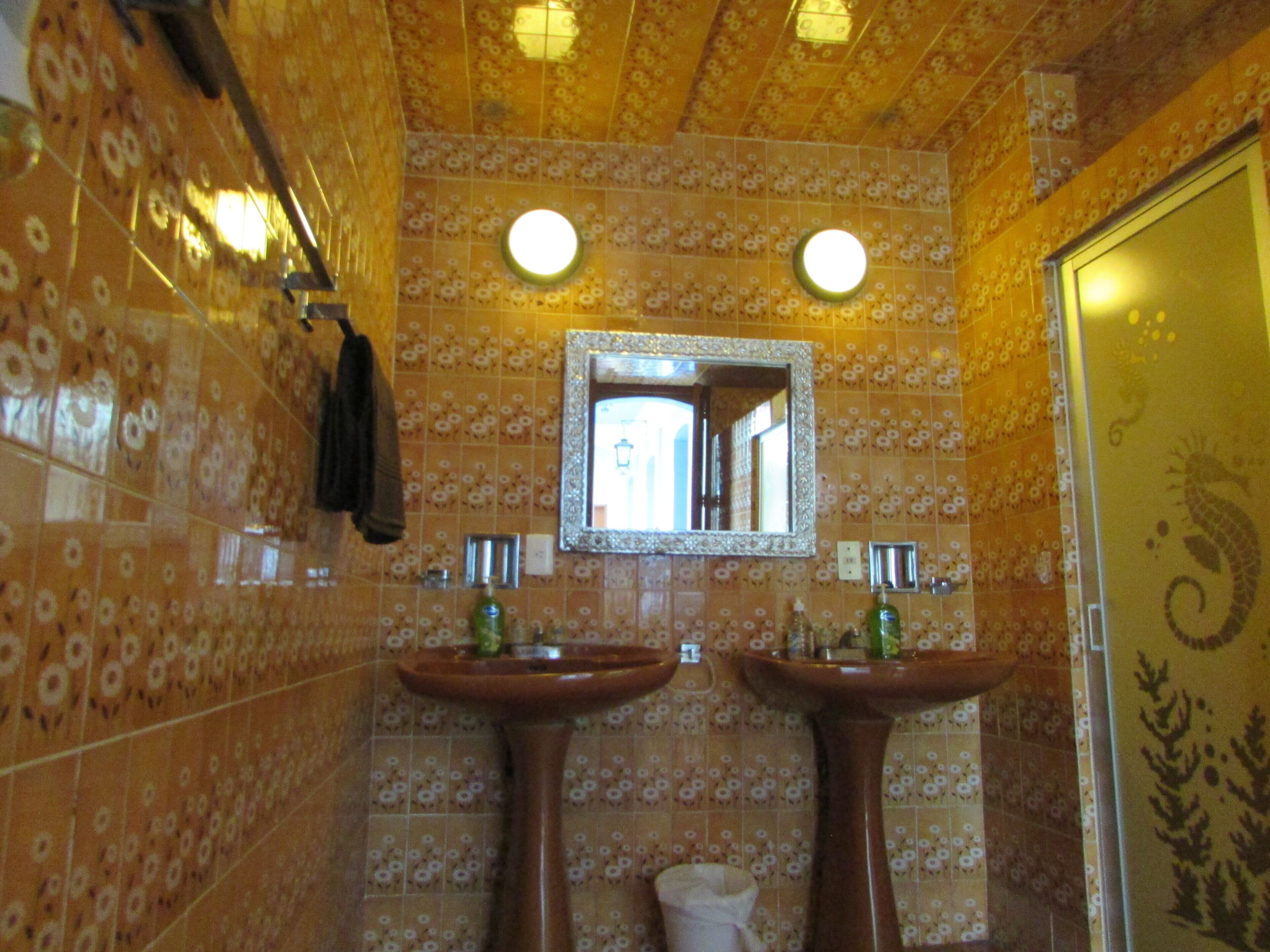 Un baño con azulejos de estampado floral, dos lavamanos, un espejo y un perchero para toallas.