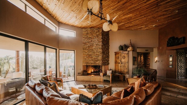 Een ruime woonkamer met een leren bank, een open haard en een groot raam met uitzicht op een terras.