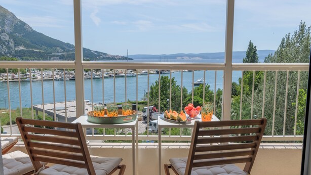 A balcony with two wooden chairs and a table with fruit and a decorative bowl. A marina with boats is visible in the background. - Beautifull attic over the sea and river coust, panoramic sea view (Omiš)