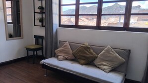 A room with a wooden floor, a sofa with cushions, a chair, a mirror, and a window with a view of a mountainous landscape. - Nice apartment in the center of Cusco (Cusco)