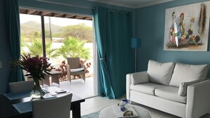 Una sala de estar con un sofá blanco, una mesa de centro redonda y un comedor con mesa y sillas. Hay un cuadro en la pared y vistas a zonas verdes desde el exterior.