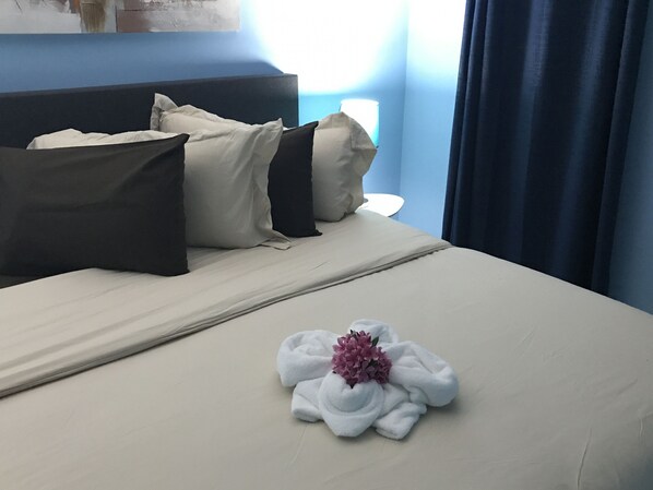 A neatly made bed with a towel and a flower, a blue wall, and a window with curtains. - Malika Apartments Blue  Westpunt Curaçao Near Beaches (willemstad)