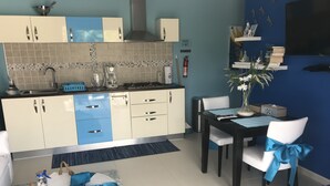 Una cocina moderna con armarios blancos y azules, una estufa a gas y una cafetera. Hay una mesa de comedor con sillas, una alfombra pequeña y una planta decorativa.