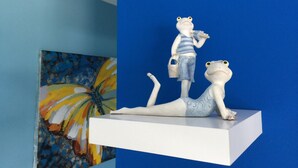 Une étagère blanche ornée de deux statuettes de grenouilles, l’une debout et l’autre allongée, le tout placé devant un mur bleu.
