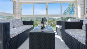 A sunlit patio with wicker furniture, a bottle of wine, and two glasses on a table, overlooking a scenic landscape.