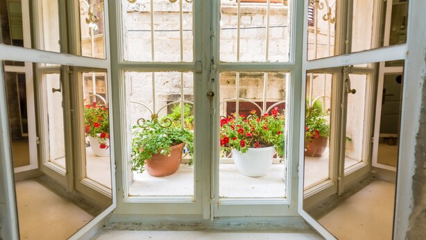 Vista a través de una ventana con marco blanco y celosías metálicas decorativas, que da a un balcón con plantas en macetas y un edificio con fachada roja al fondo.