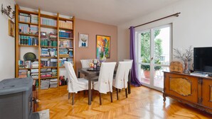 Een eetgedeelte met een houten tafel, witte stoelen, een boekenplank vol boeken, een schilderij aan de muur en een raam met gordijnen.