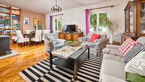 Een woonkamer met een houten visgraatvloer, een zwart-wit gestreept tapijt, een eethoek met stoelen en een tafel, een boekenkast, een televisie en een kast met glazen deuren.