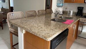 A kitchen with granite countertops, a dishwasher, a sink, a coffee maker, and wicker chairs. - Greate apartment at Nuevo Vallarta (Las Jarretaderas)