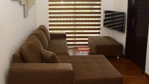 A living room with a brown sectional sofa, a small ottoman, a flat-screen TV mounted on the wall, and a window with horizontal blinds. - MARINERS CABIN LUXURY HOLIDAY APARTMENT  (Nuwara Eliya)