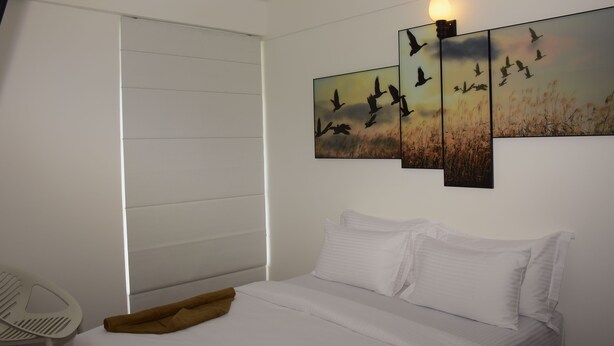 A bedroom with a bed, white bedding, a white chair, and a wall-mounted artwork of birds flying. - MARINERS CABIN LUXURY HOLIDAY APARTMENT  (Nuwara Eliya)