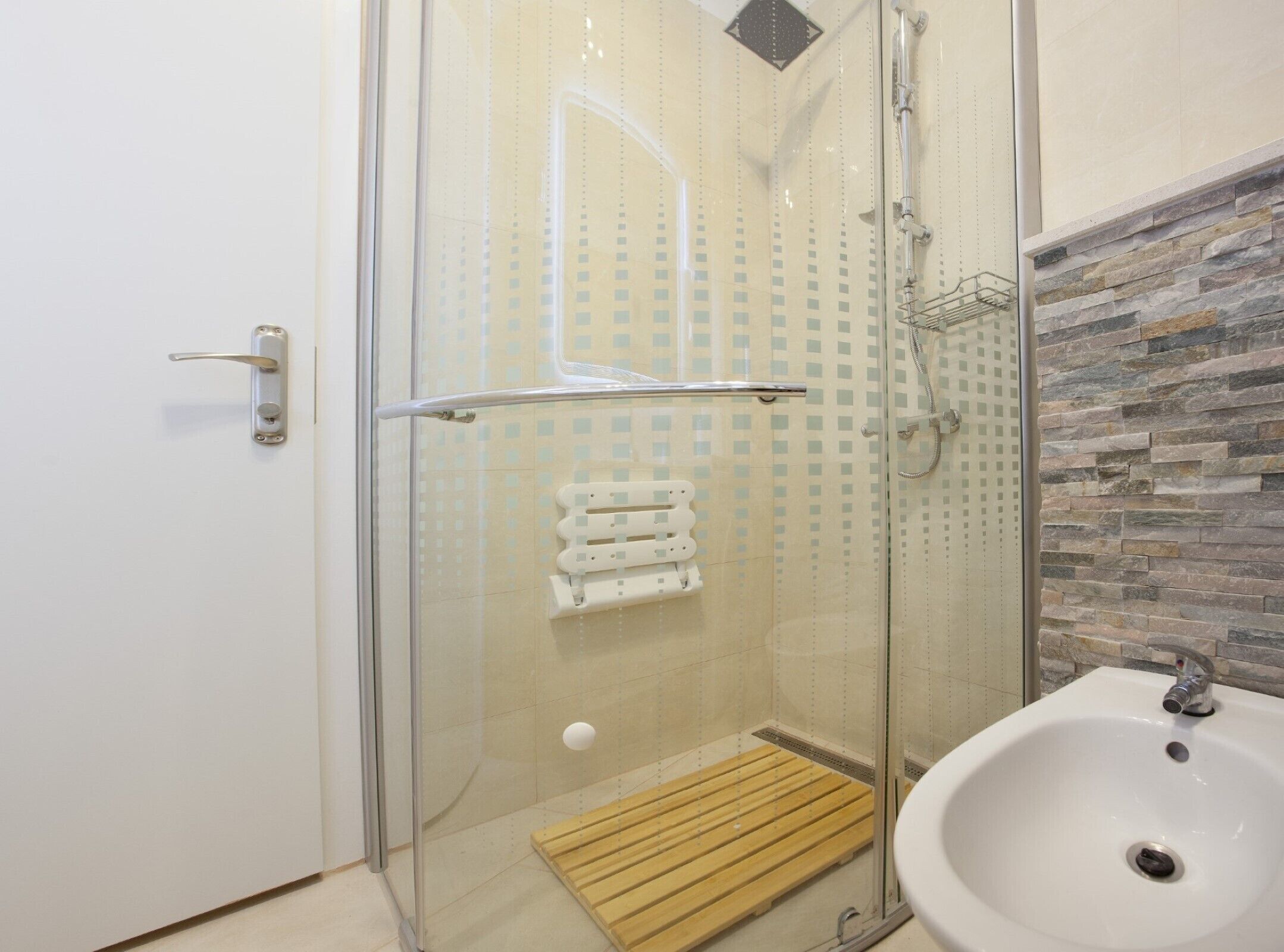 A glass-enclosed shower with a wooden mat, a white sink, and a stone-textured wall.
