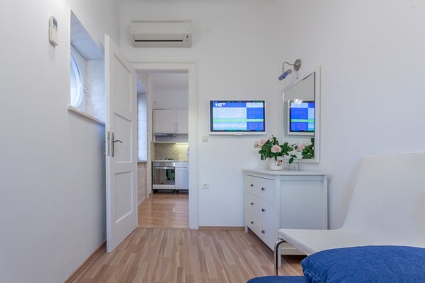 A modern, well-lit room with a wooden floor, a white bed, a small desk with a computer monitor, and a white dresser.