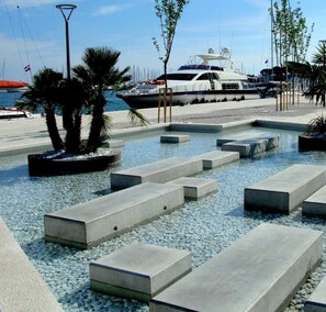 A waterfront promenade with concrete steps leading to a pool, surrounded by palm trees and boats in the background.